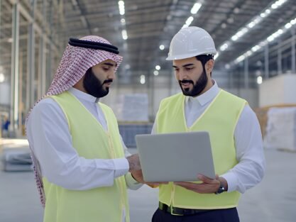 Two men in hard hats and safety vests looking at a laptop in a factory setting.